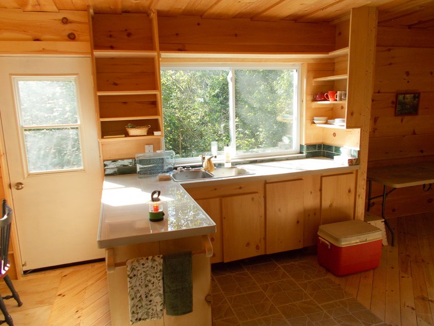 Large window lights L-shaped kitchen with double sink. Smooth white tile countertop with solid wood cabinents below and solid wood shelves above.