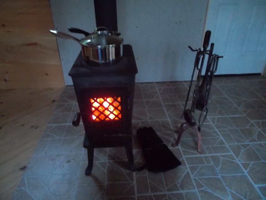 Close up of woodstove with warm glow and two pots on top. Nearby are fire gloves and fire tools